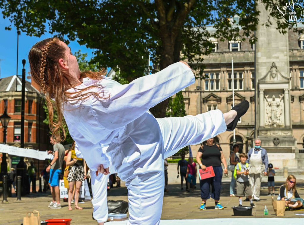 Image of Alissa in a white boiler suit leaning backword with one leg raised in the air.